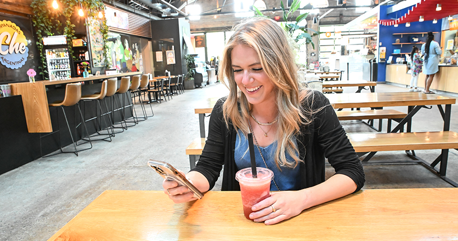 A person smiling at their phone while drinking a smoothie at a local shop.