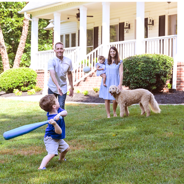 Young boy playing baseball with dad with mom and little sister and dog in yard in front of a house.