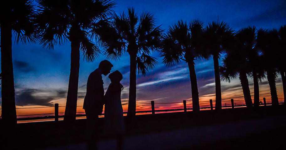 A couple standing romantically under palm trees at sunset.